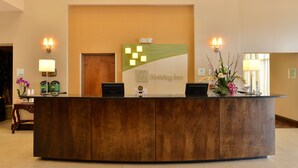 A reception desk with a wooden finish, a green and white logo, and two wall-mounted lamps. - Holiday Inn Montgomery Airport South by IHG (Montgomery)
