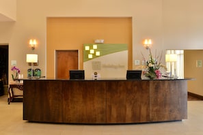 A reception desk with a wooden finish, a green and white logo, and two wall-mounted lamps. - Holiday Inn Montgomery Airport South by IHG (Montgomery)