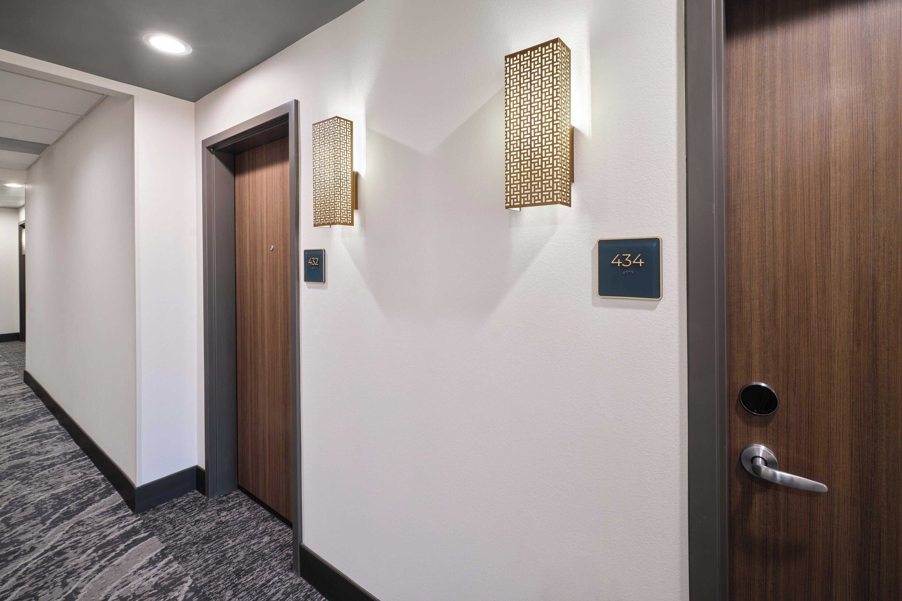 A hotel hallway with a carpeted floor, two doors with numbers 432 and 433, and wall-mounted light fixtures.