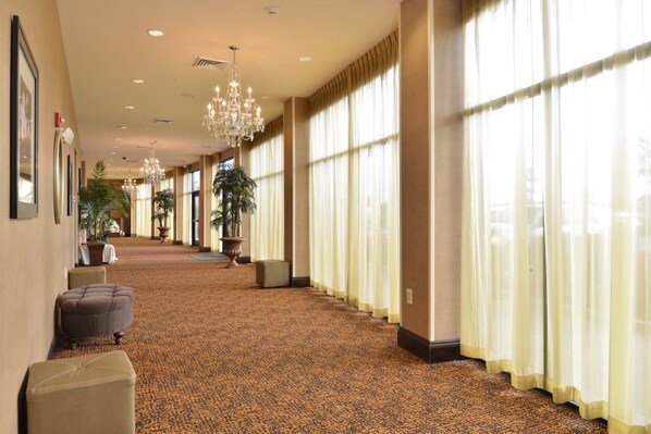 A hotel corridor with a patterned carpet, large windows with sheer curtains, and a chandelier.