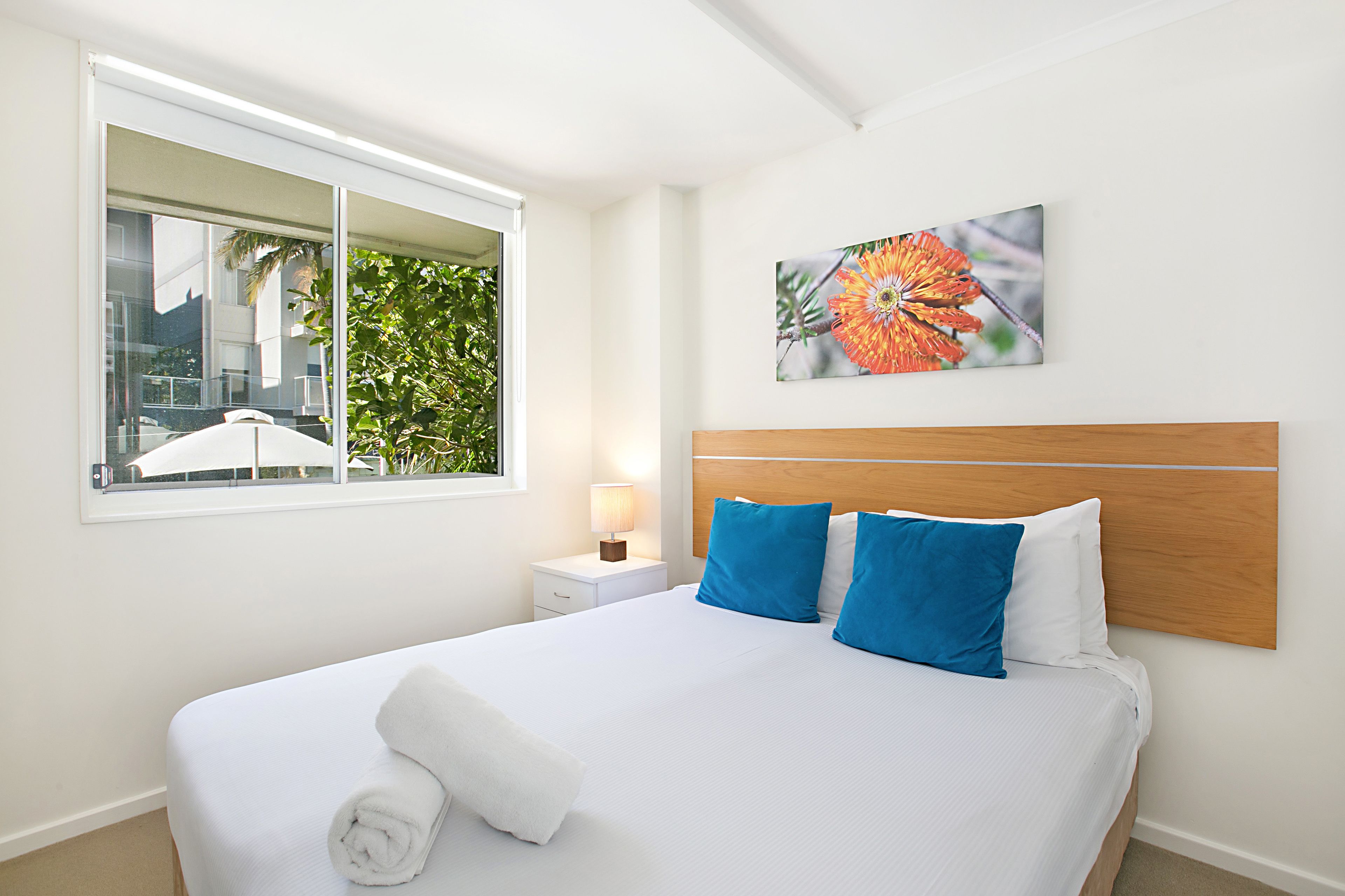 Una cama bien hecha con almohadillas blancas y azules, una mesita de noche blanca con una lámpara y una ventana con vistas a zonas verdes y edificios.
