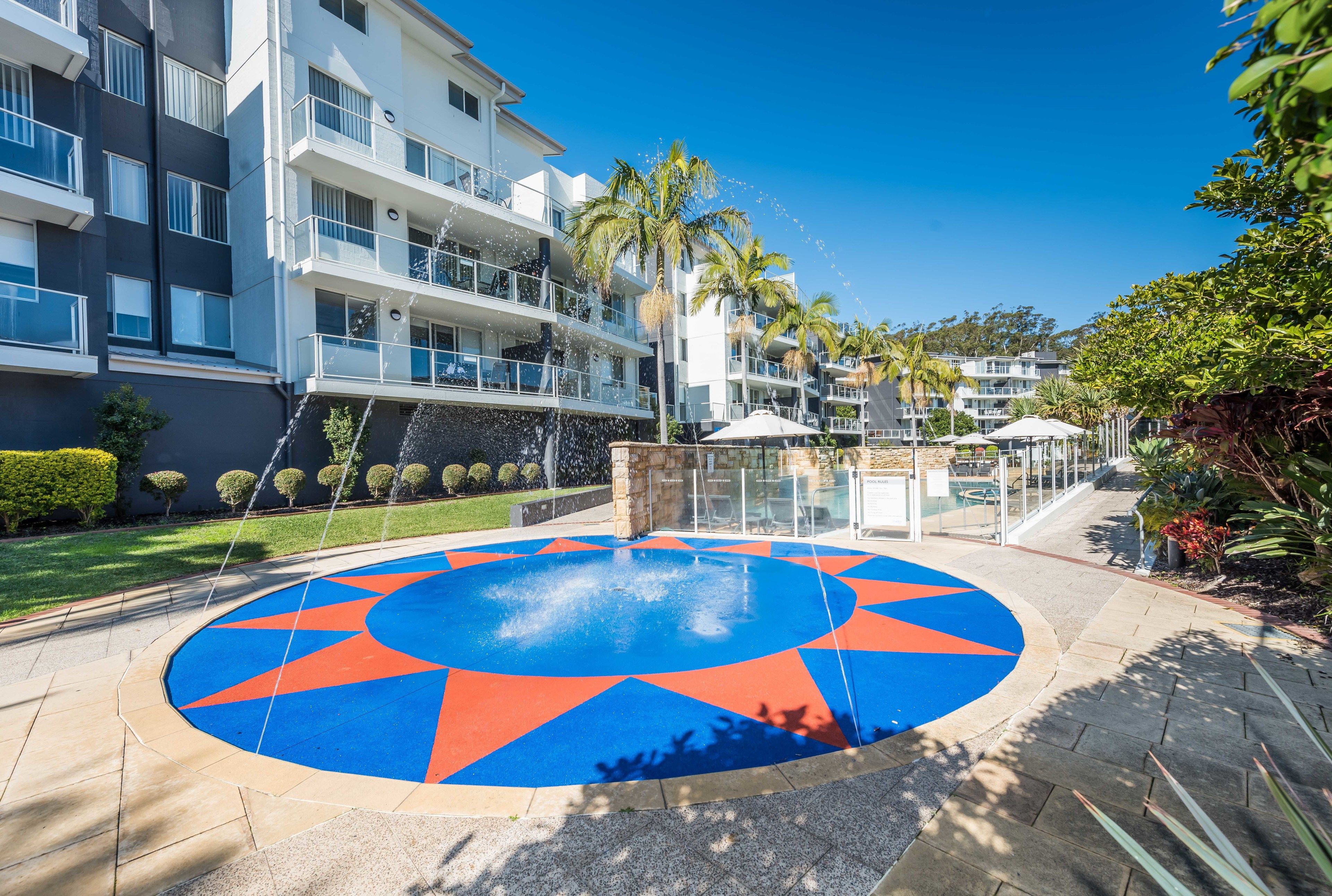 Un patio con fuente circular, palmeras y edificios de apartamentos modernos.