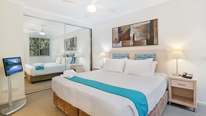 Una habitación de hotel con una cama grande, un televisor de pantalla plana, un ventilador de techo, una mesita de noche con lámpara y un cuadro en la pared.