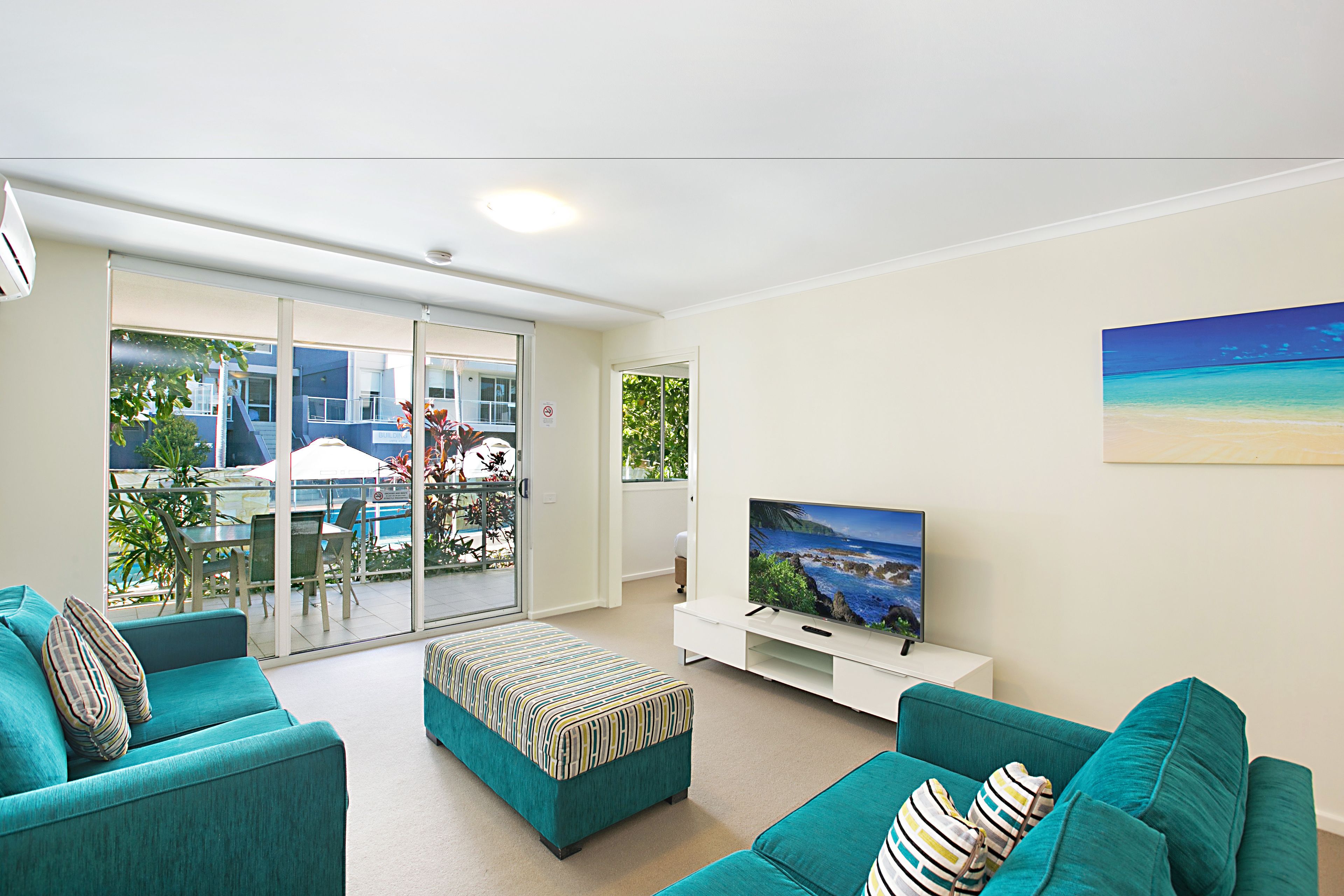 A modern living room with teal sofas, a flat-screen TV, and a sliding glass door leading to an outdoor area with a pool and patio furniture.