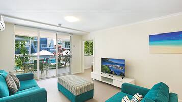 A modern living room with teal sofas, a flat-screen TV, and a sliding glass door leading to an outdoor area with a pool and patio furniture.