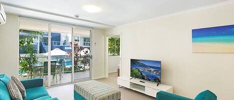 A modern living room with teal sofas, a flat-screen TV, and a sliding glass door leading to an outdoor area with a pool and patio furniture.