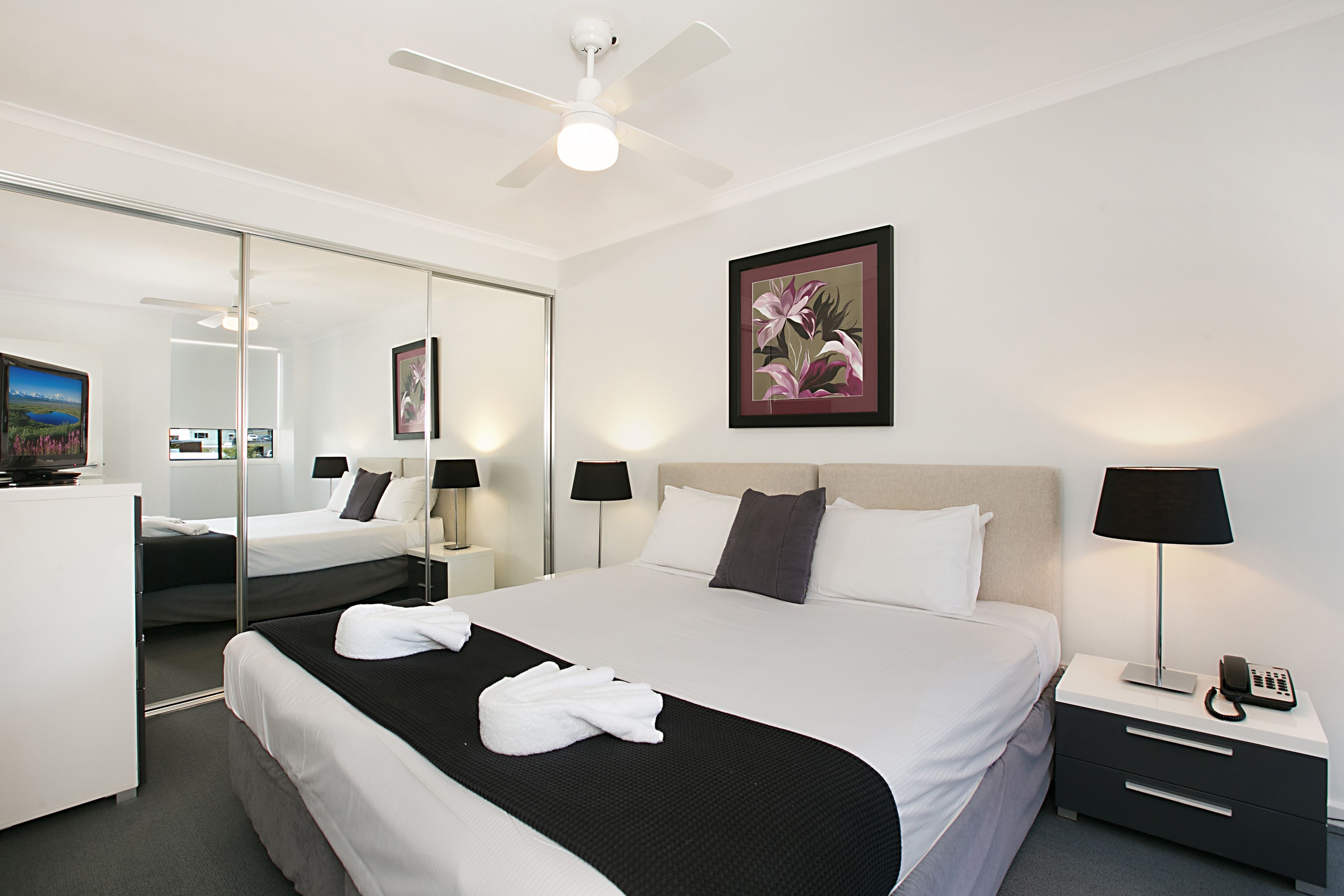 Une chambre d’hôtel avec un grand lit, des tables de chevet, un ventilateur de plafond, une œuvre d’art florale encadrée et un téléviseur.