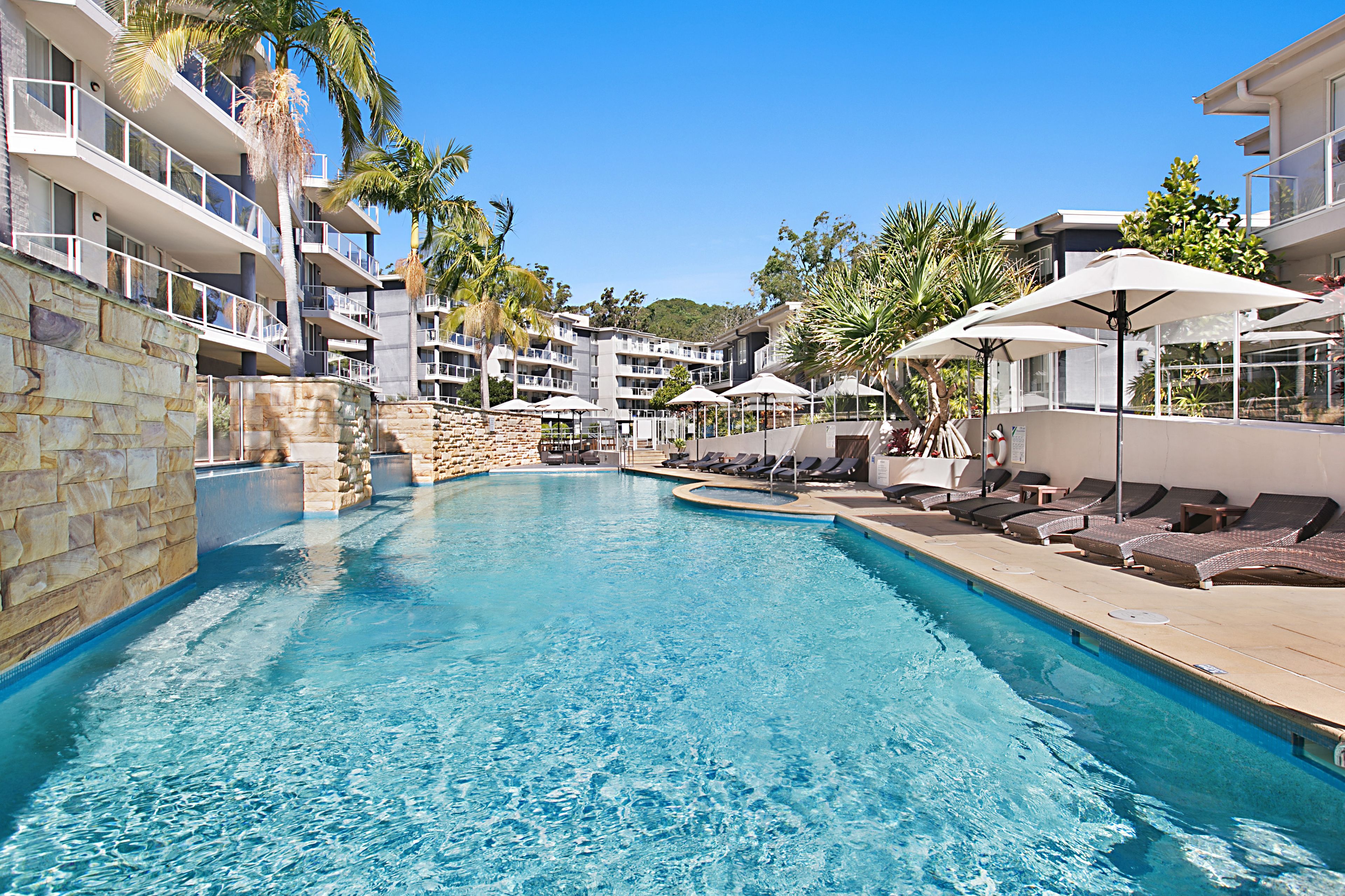 A swimming pool with lounge chairs and umbrellas, surrounded by stone walls and palm trees.