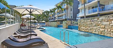 A poolside area with lounge chairs and umbrellas, a stone wall, and a clear blue sky.