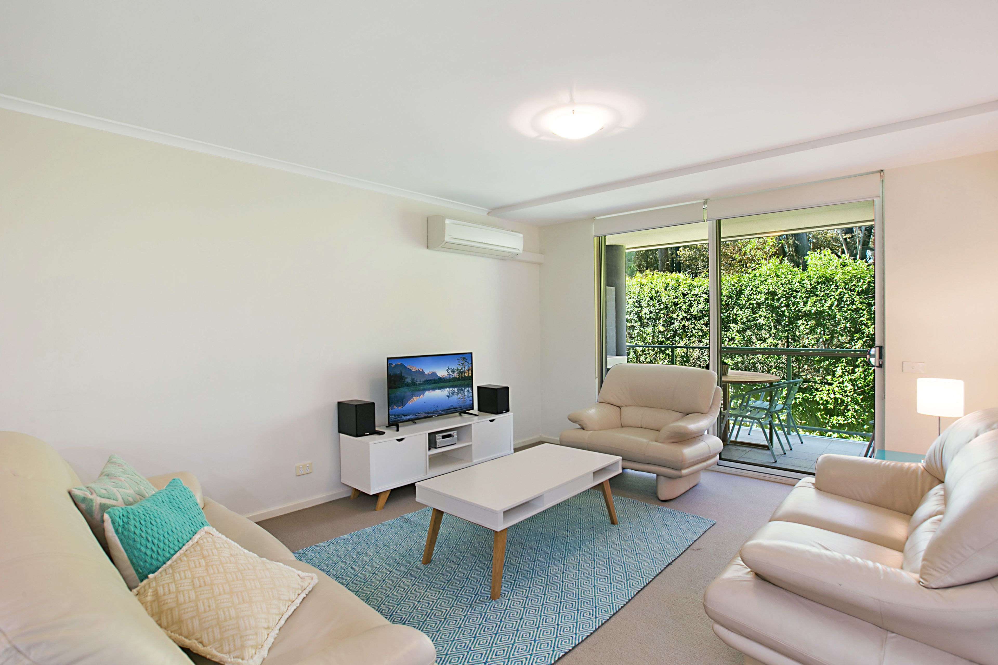 A modern living room with a flat-screen TV, white sofas, a coffee table, and a balcony with a view of greenery.