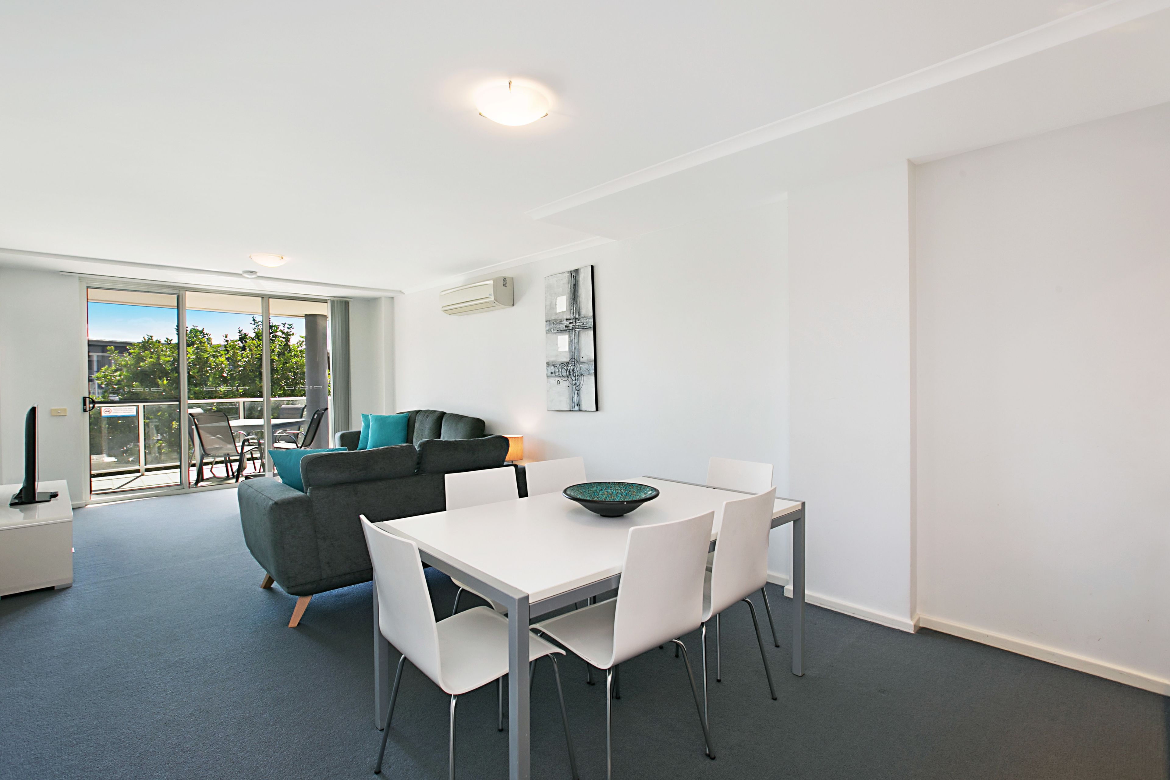 A modern dining area with a white table and chairs, a grey sofa, and a glass door leading to an outdoor balcony.
