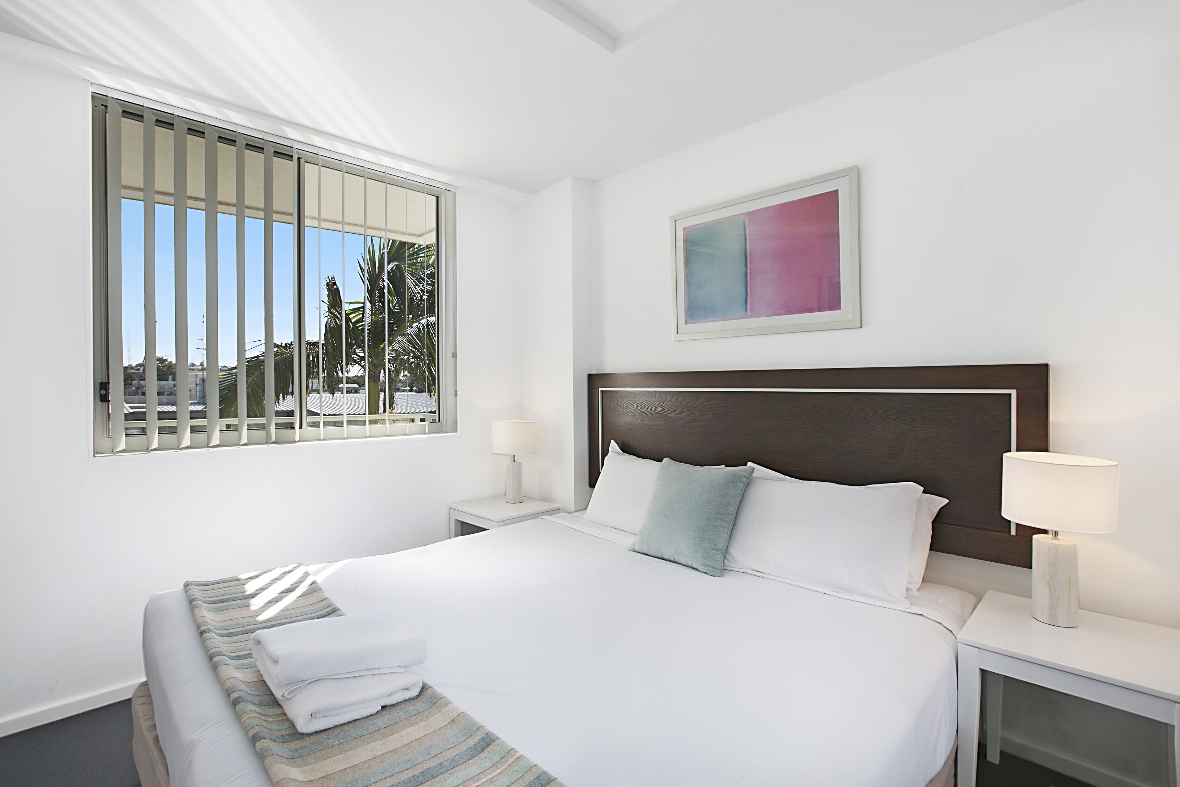 Una cama bien hecha con sábanas blancas y una manta a rayas, una mesita de noche con una lámpara y una ventana con persianas.