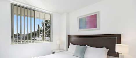 Una cama bien hecha con sábanas blancas y una manta a rayas, una mesita de noche con una lámpara y una ventana con persianas.