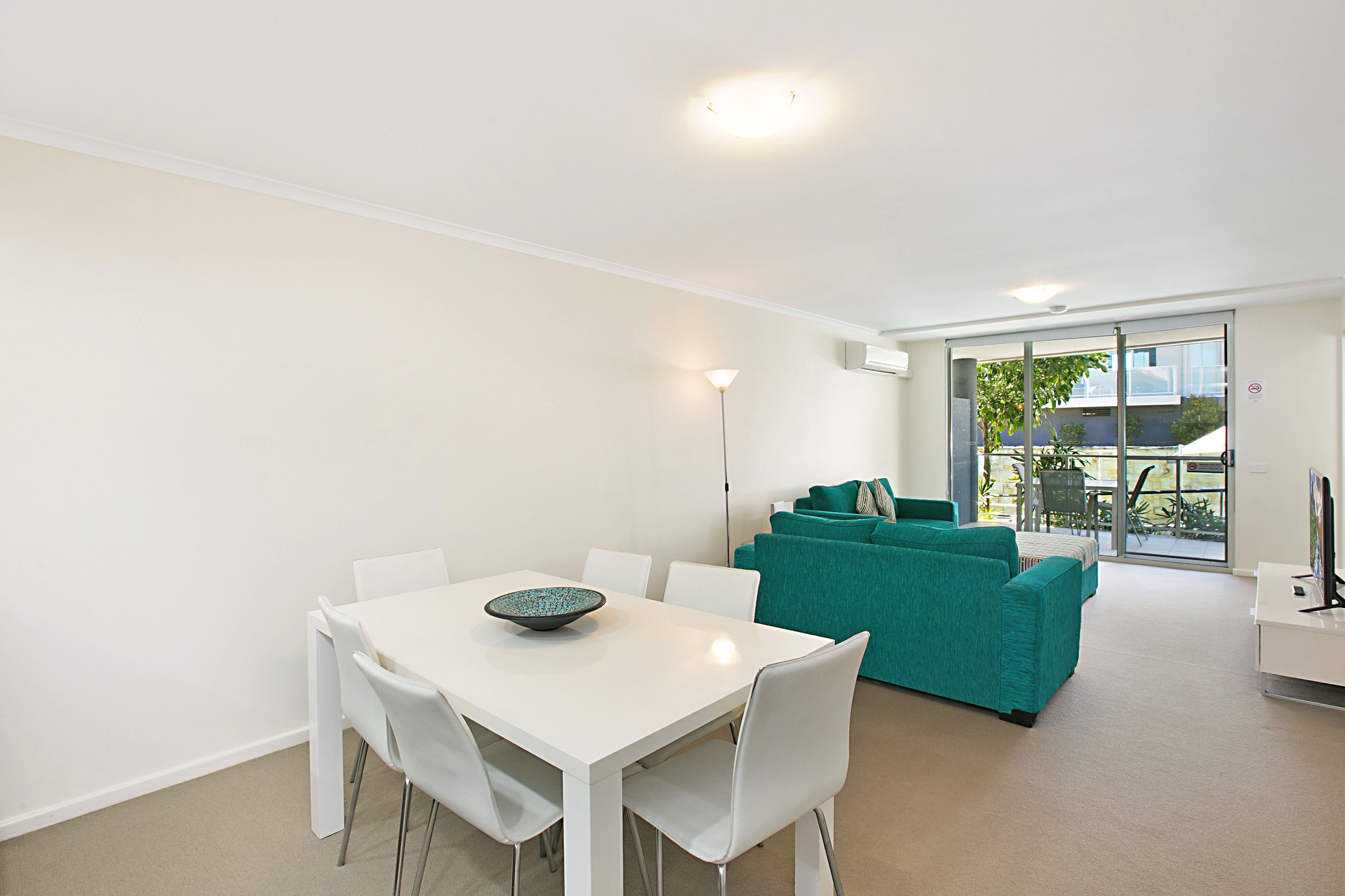 A modern living room with a white dining table, teal sofa, and a large window offering a view of the outdoors.