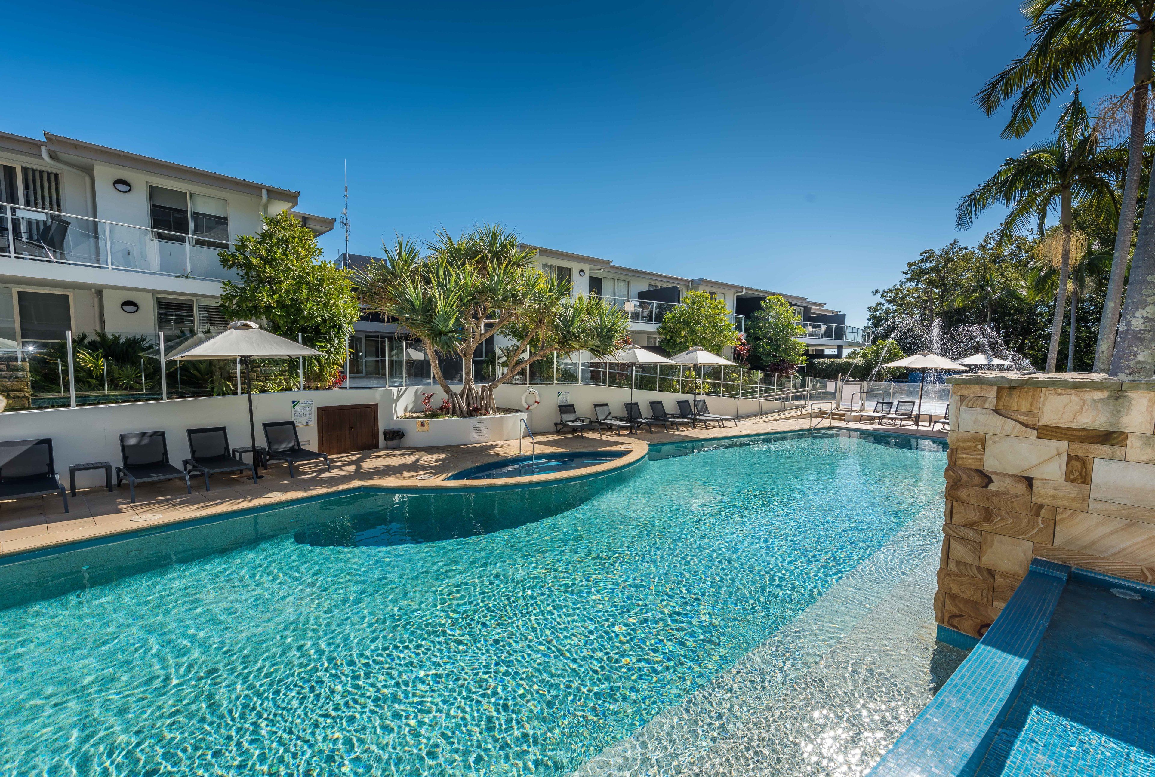 A swimming pool with lounge chairs and umbrellas, surrounded by palm trees and a building with balconies.