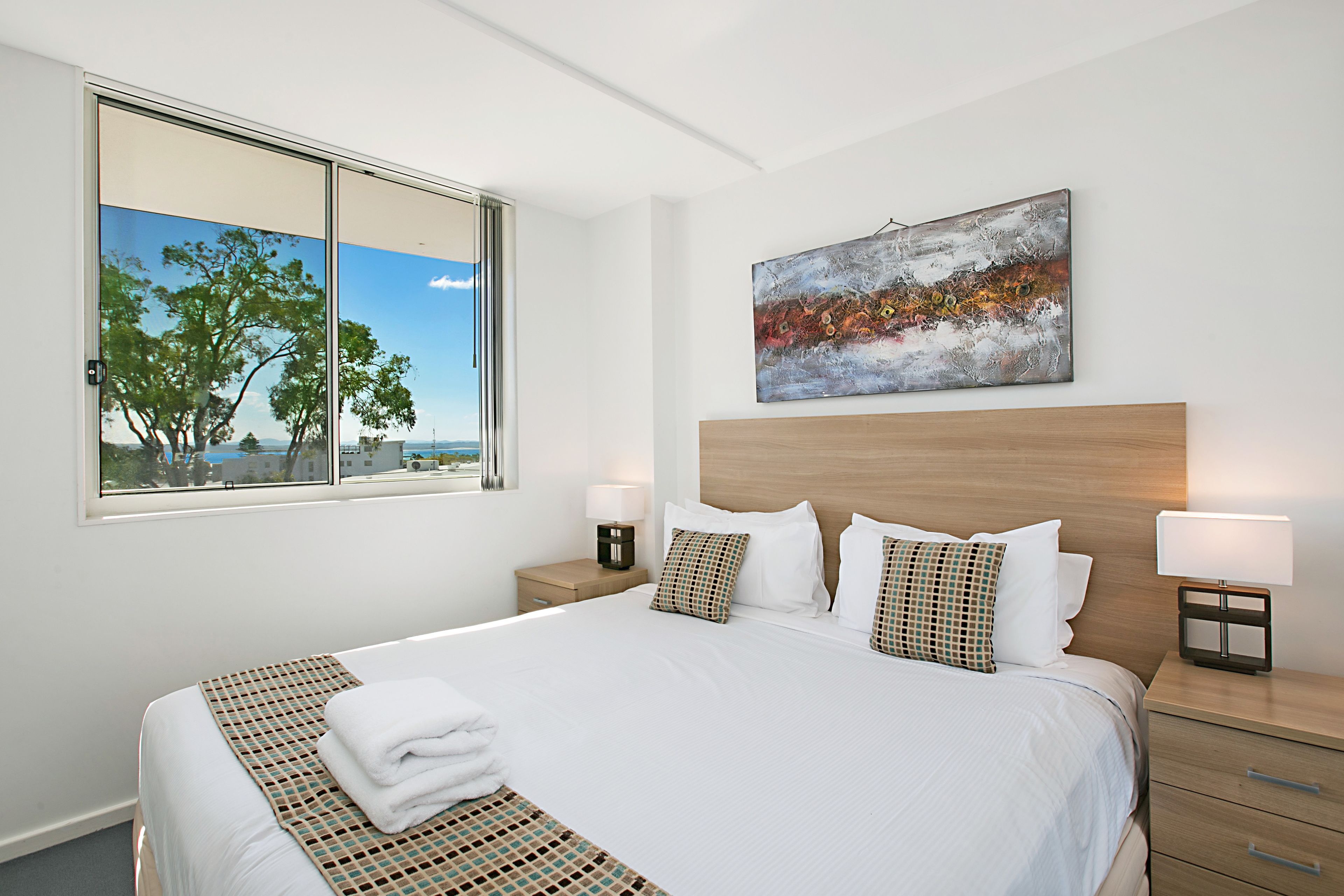 Habitación de hotel con una cama grande, vistas a árboles y una masa de agua a través de una ventana, un cuadro en la pared y una mesita de noche con una lámpara.