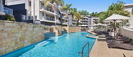 A swimming pool with lounge chairs and umbrellas, surrounded by multi-story buildings with balconies.