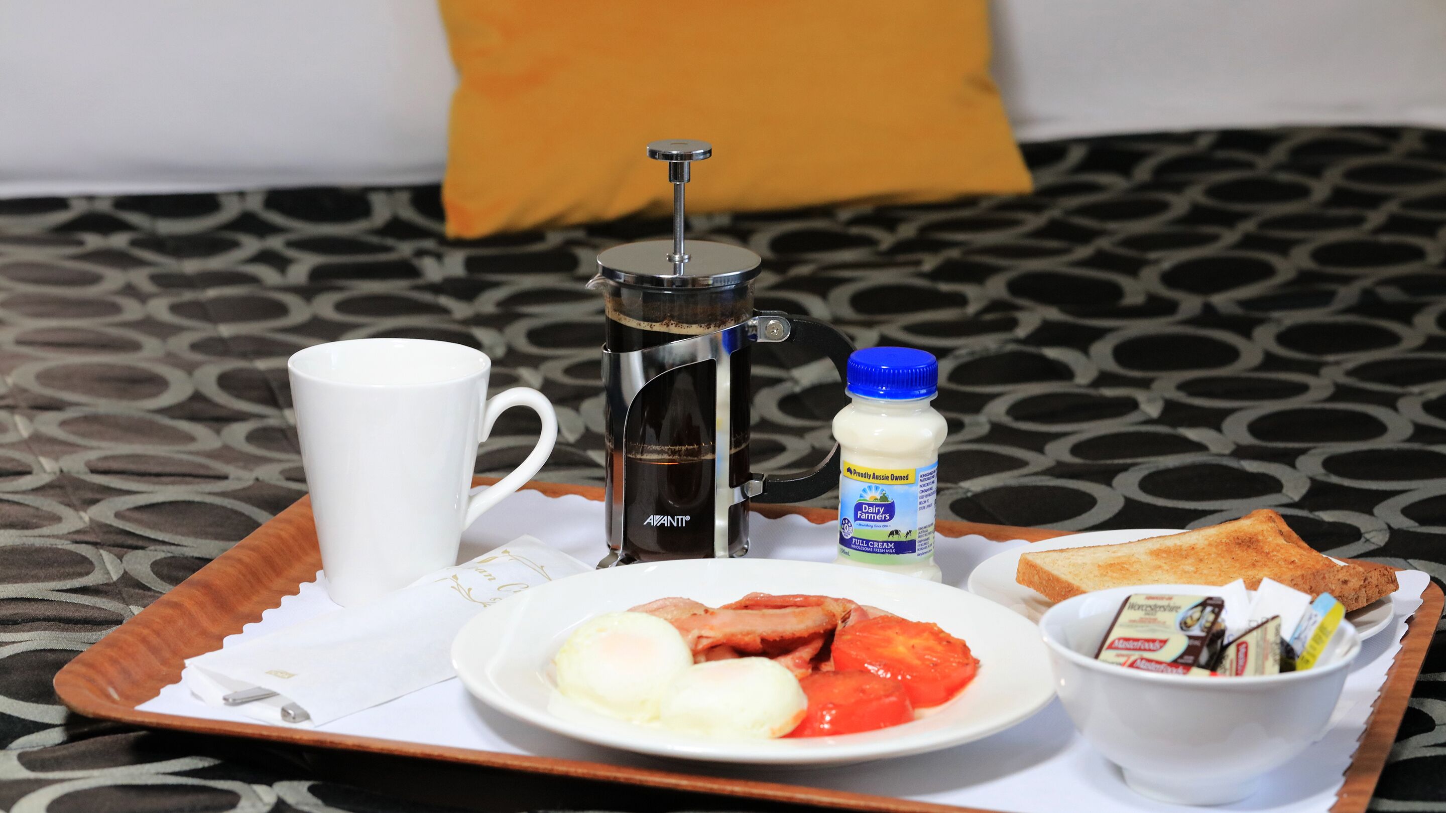 Ett frukostbricka med en kaffebryggare, en flaska mjölk, rostat bröd, ägg och en kopp placerat på ett mönstrat överkast.