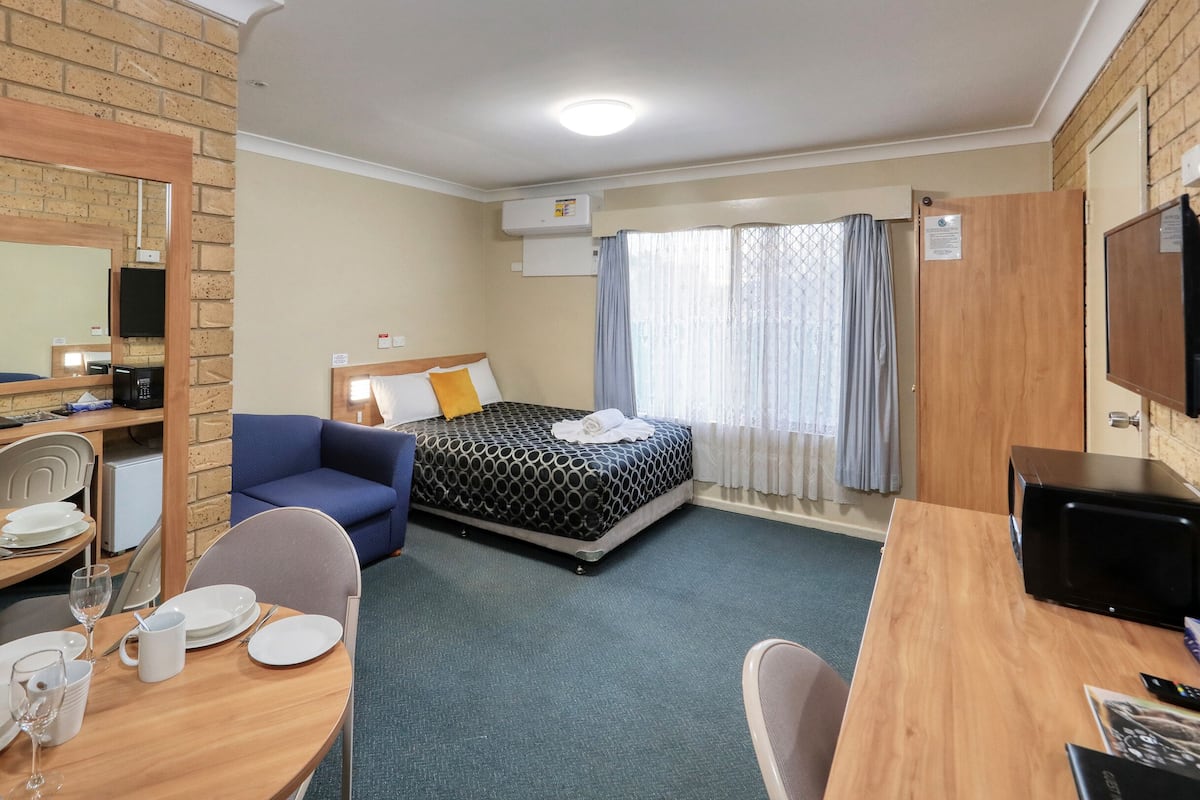 A hotel room with a bed, a desk with a chair, a dining table set for two, a TV, and a small kitchenette.