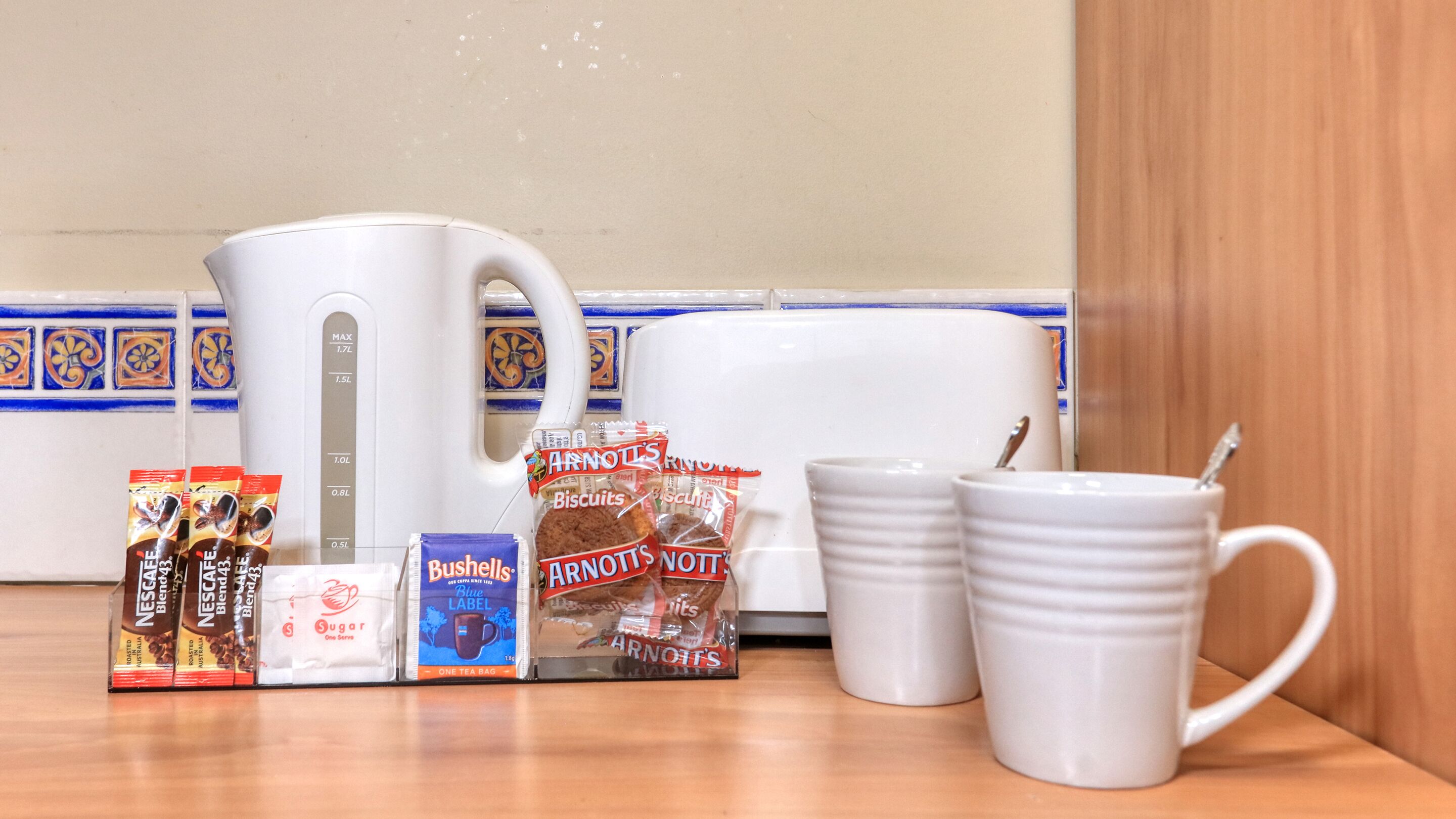 A kitchen counter with a white coffee maker, two white mugs, and various food items including biscuits and coffee blends.