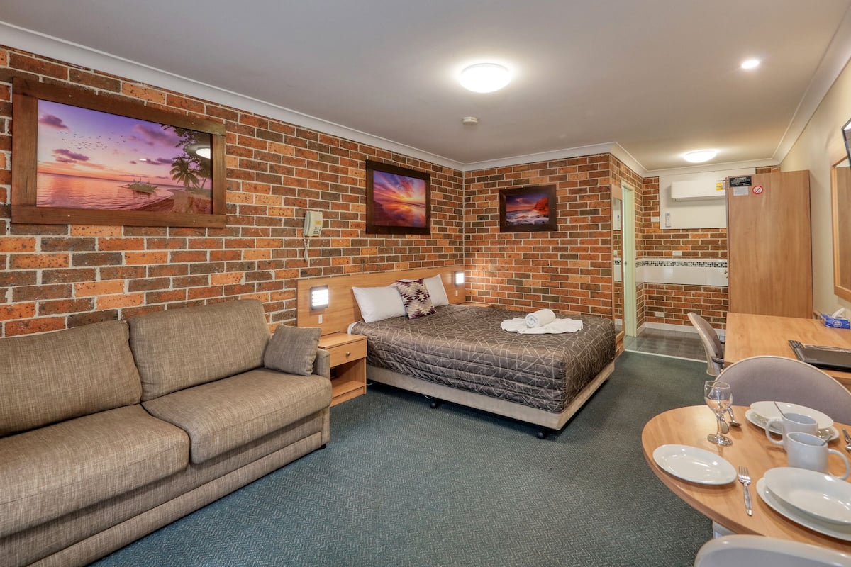 A hotel room with a brick wall, a bed with a quilt, a sofa, a dining table set with plates and glasses, and a TV mounted on the wall.