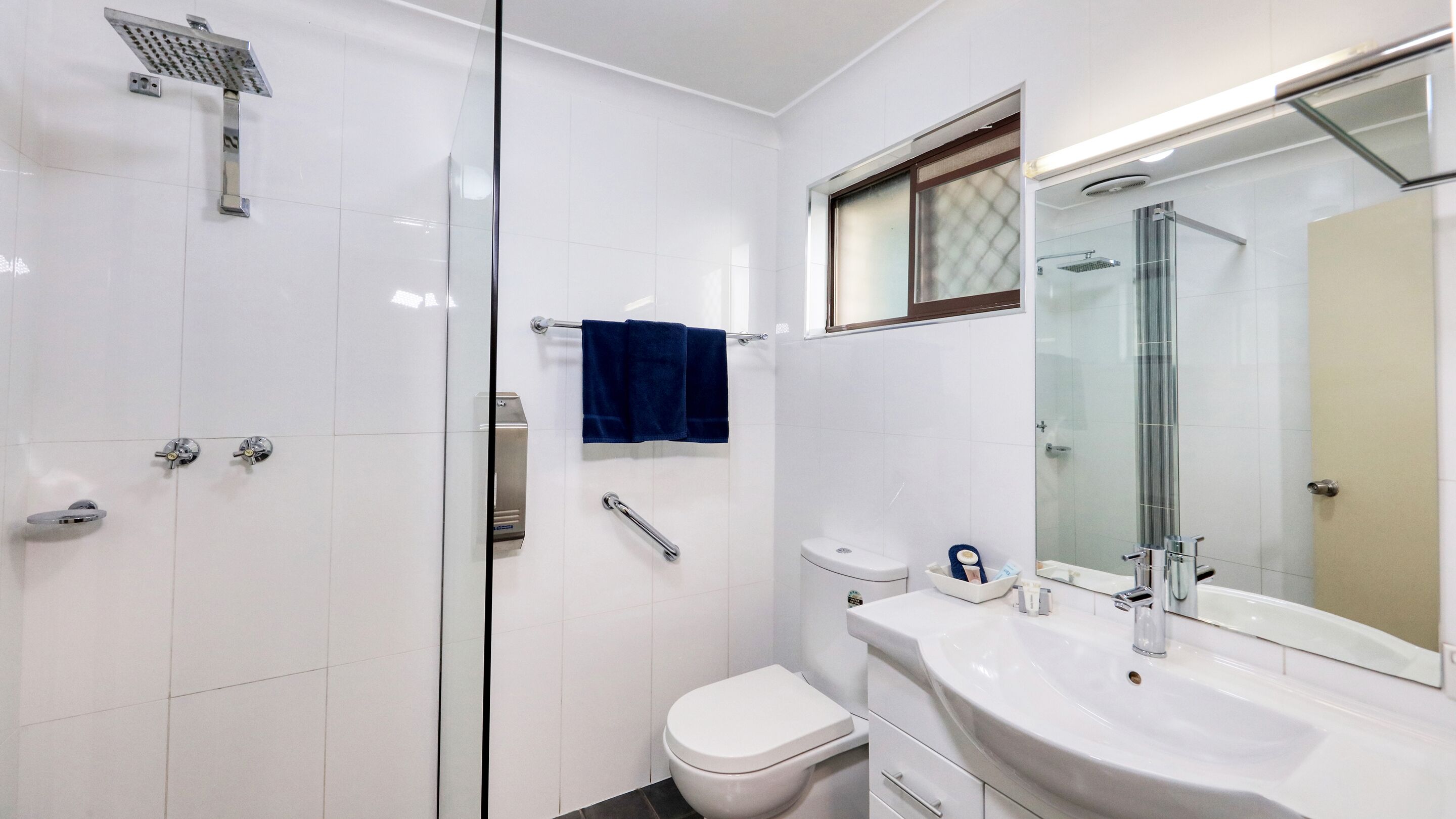 Un baño moderno con cabina de ducha de vidrio, inodoro, lavamanos con espejo y ventana sobre el lavamanos.