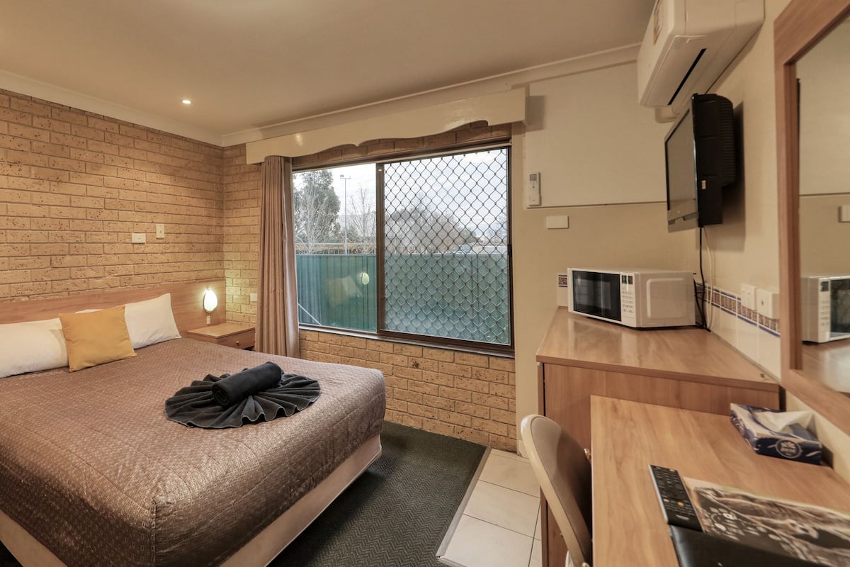 A hotel room with a brick wall, a bed with a brown blanket, a desk with a microwave, and a TV on the wall.