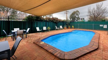 Una piscina rettangolare con bordo in mattoni, circondata da posti a sedere all'aperto e una recinzione verde.