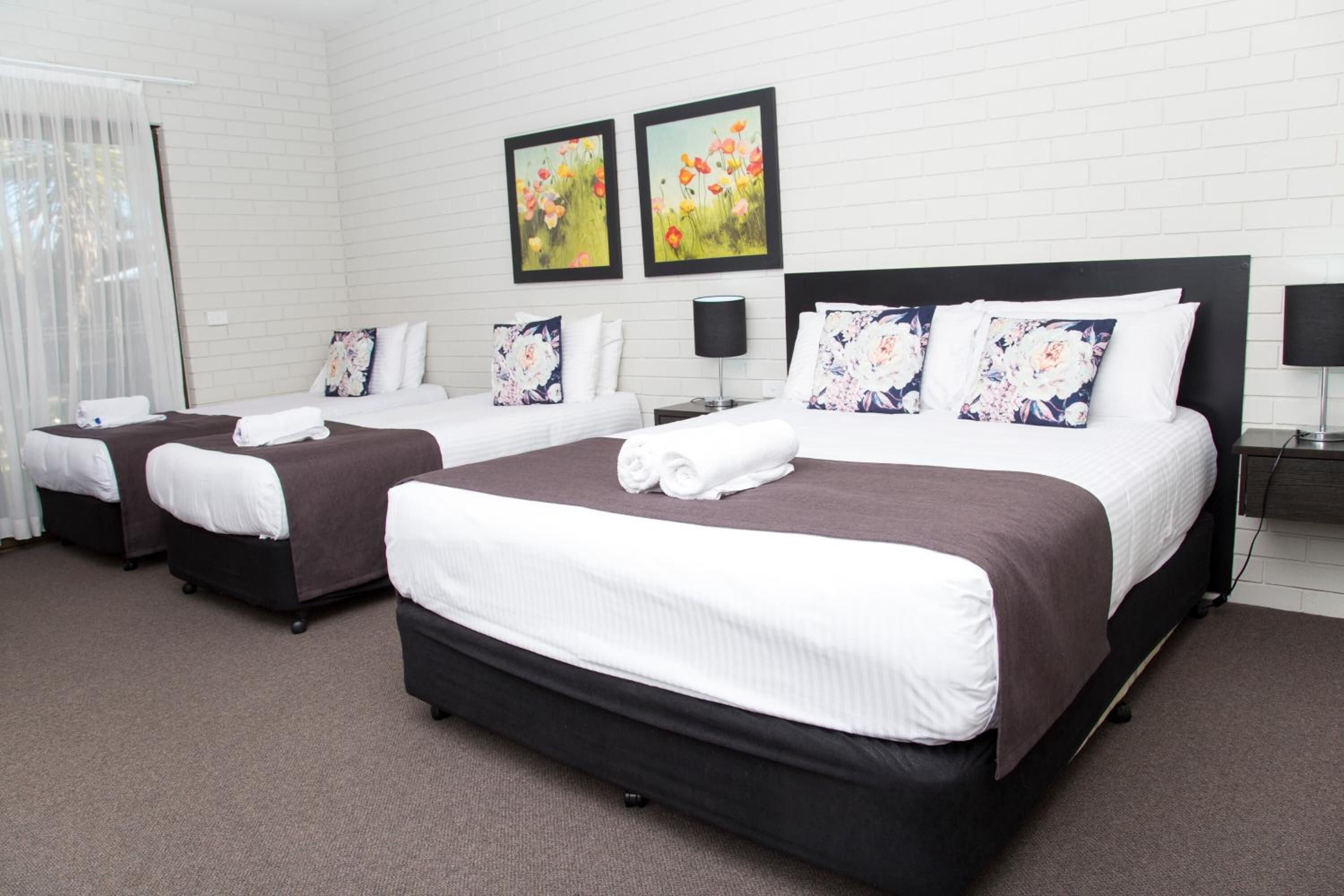 A hotel room with three beds, each with a brown headboard and white bedding. There are two framed floral paintings on the wall above the beds.