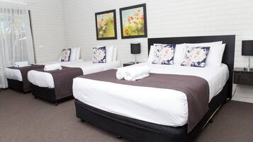 A hotel room with three beds, each with a brown headboard and white bedding. There are two framed floral paintings on the wall above the beds.