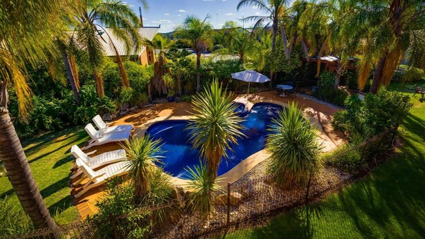 A swimming pool surrounded by lush greenery and palm trees, with a clear blue sky above. - Crystal Fountain Motel Albury (Lavington)