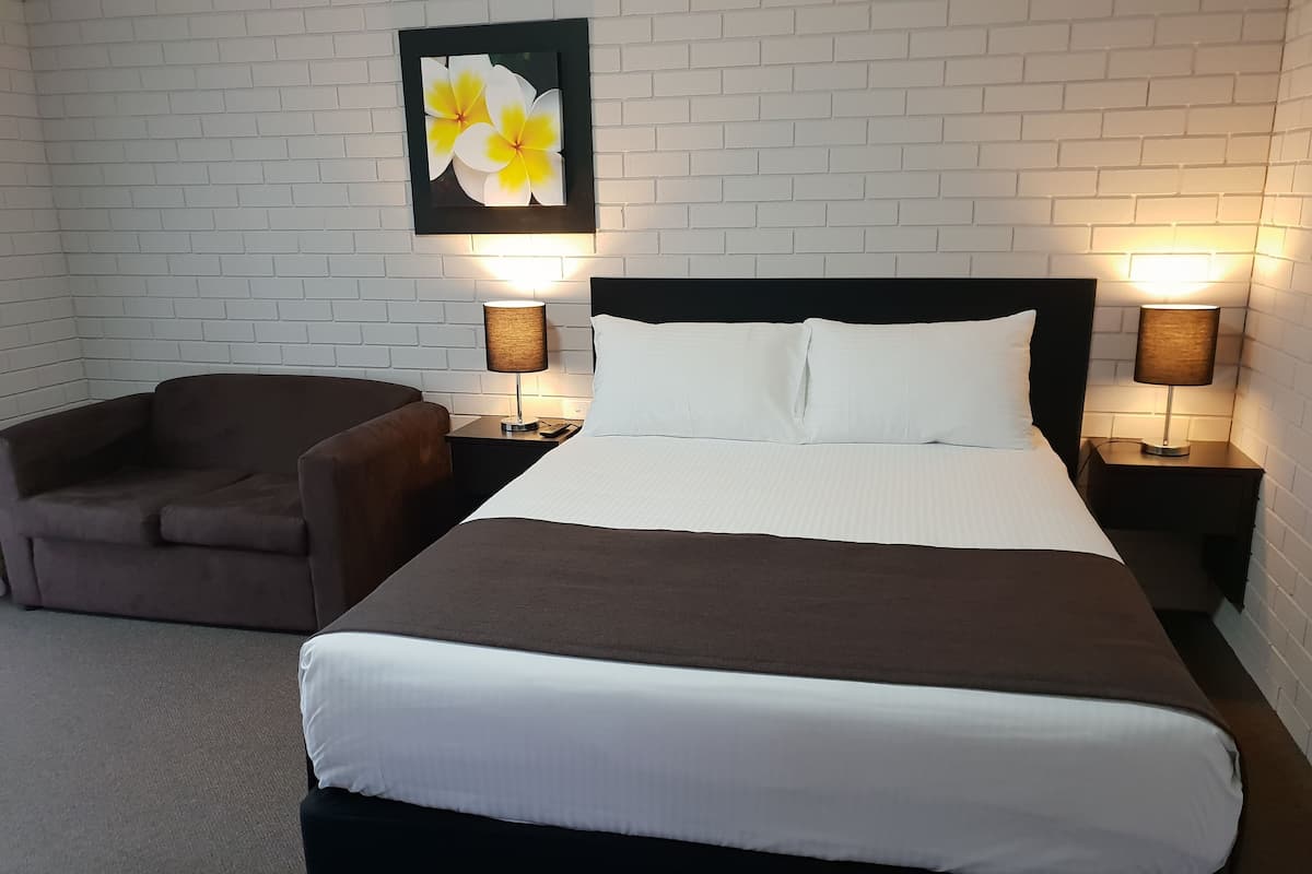 A hotel room with a large bed, two bedside tables with lamps, a brown armchair, and a white brick wall with a floral painting.