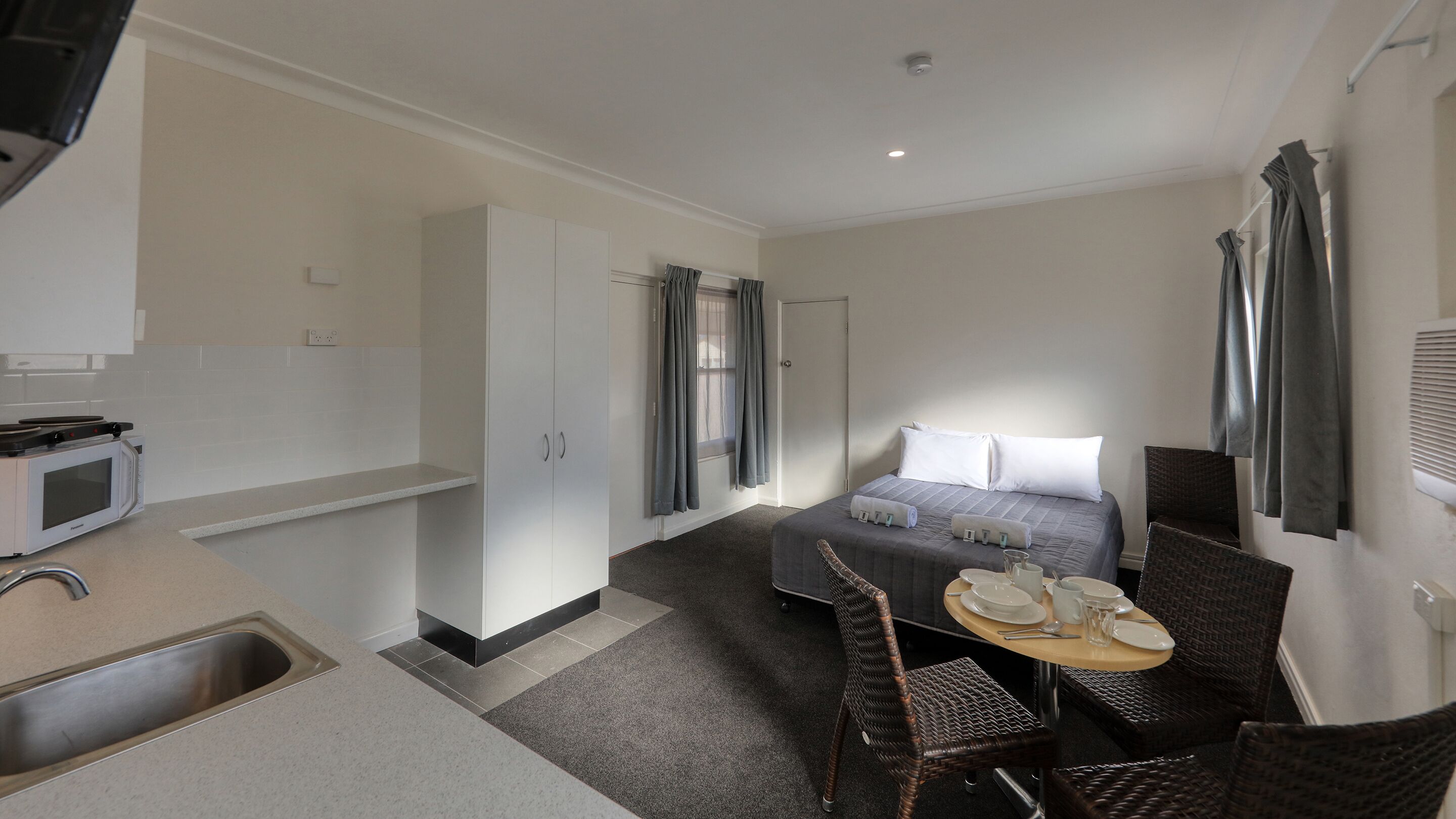 A compact hotel room with a kitchenette, a bed with gray bedding, a dining area with a table set for two, and a bathroom with a shower and toilet.