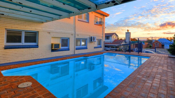 A swimming pool with a brick deck, a covered patio, and a view of a residential area with houses and a clear sky.