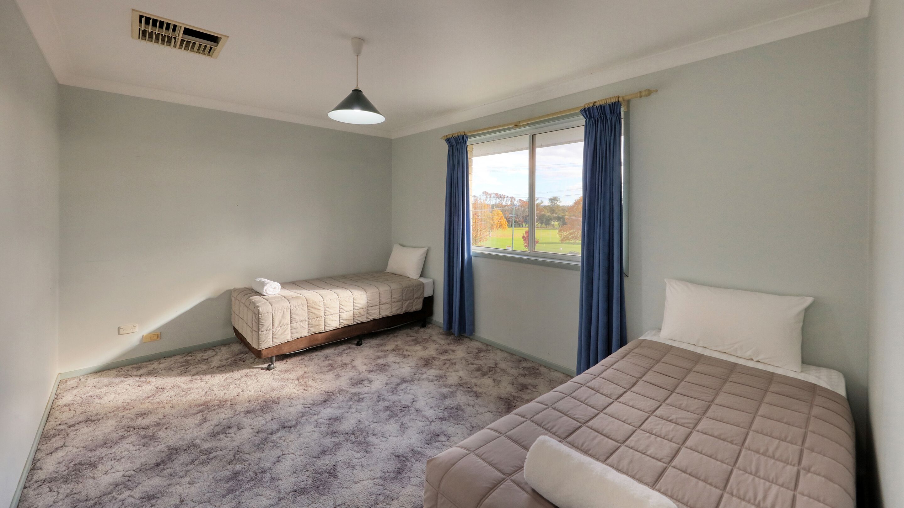 A bedroom with two beds, a window with curtains, a ceiling vent, and a light fixture.
