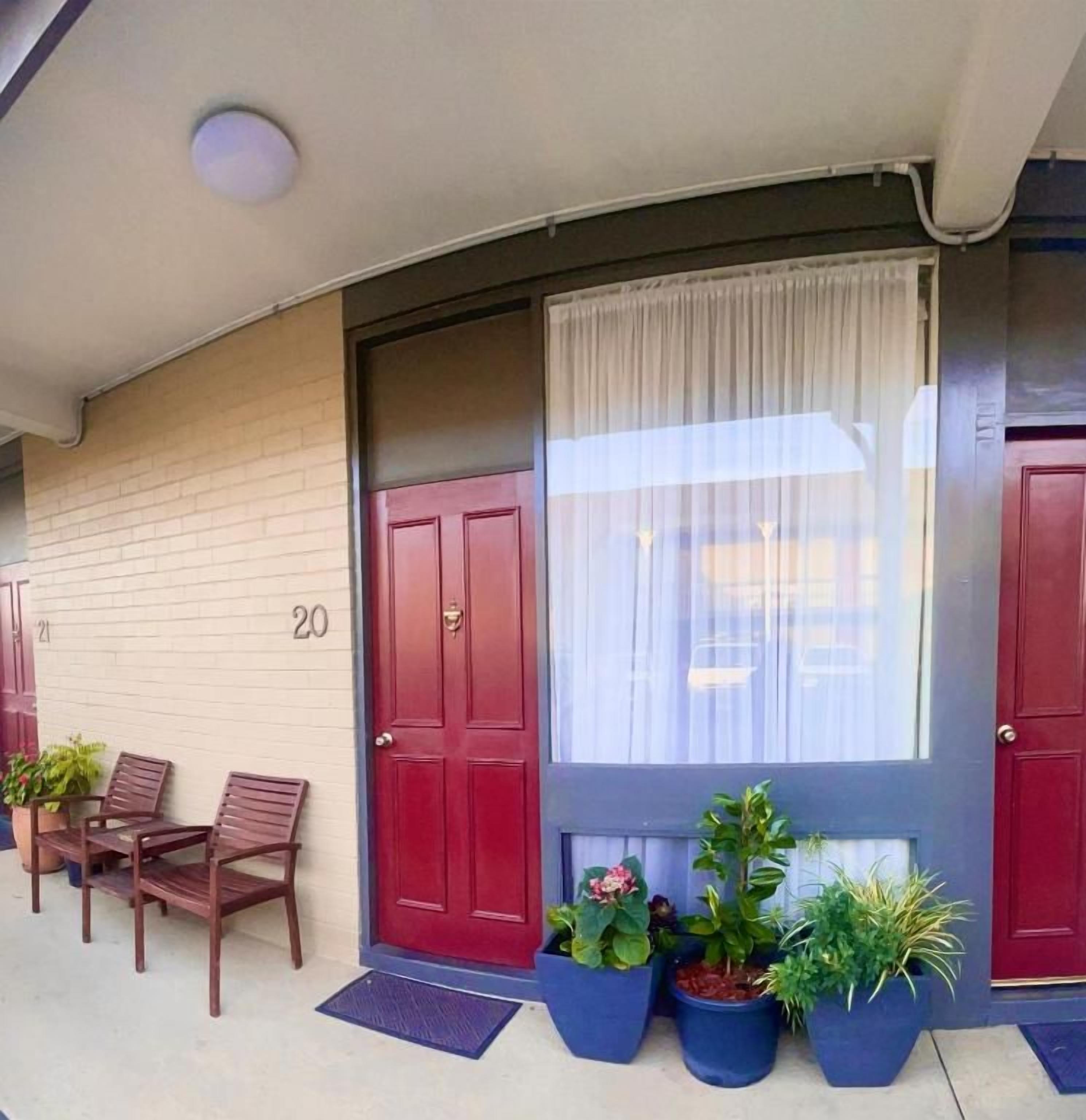 A building entrance with a red door, a window with a white curtain, and potted plants.