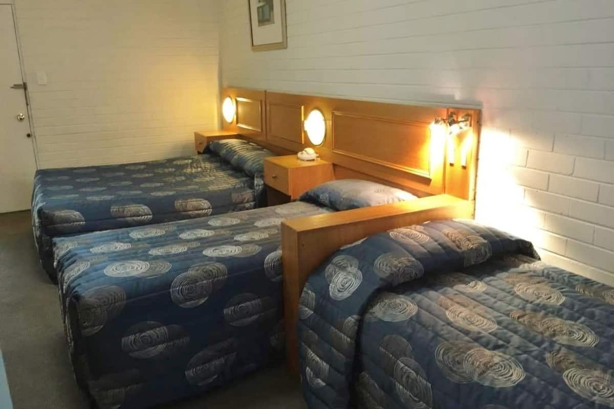 A room with three beds, each with a blue patterned duvet and wooden headboards. There are bedside lamps and a small table between the beds.
