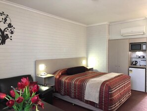 Une chambre d’hôtel comprenant un lit, un bureau, une chaise, une table de chevet, un climatiseur mural et une kitchenette équipée d’un four à micro-ondes et d’un réfrigérateur.