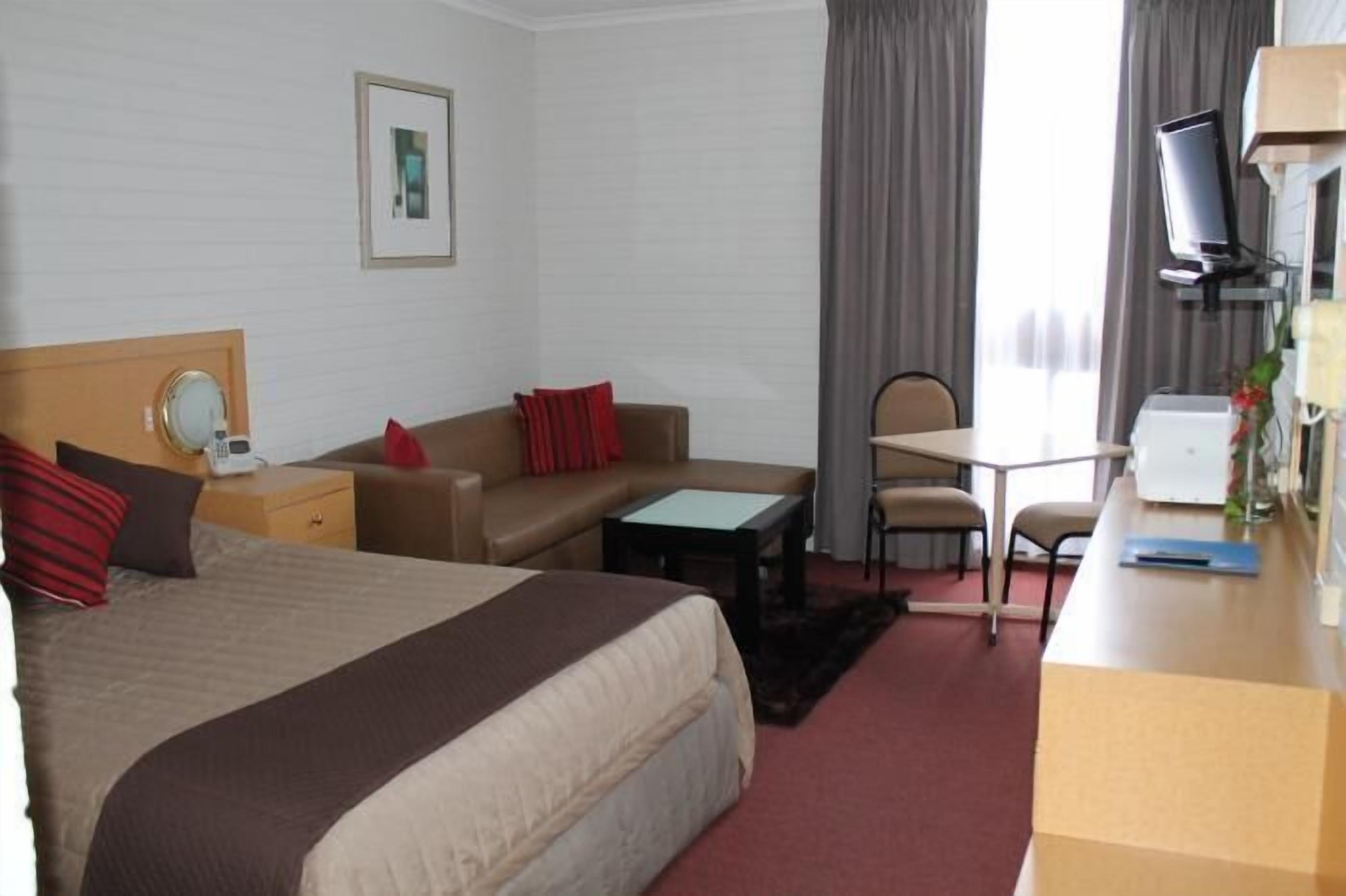 A hotel room with a bed, a desk, a chair, a small table, a mirror, and a television.
