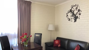 Une pièce comprenant une table à manger, deux chaises, un canapé en cuir noir avec des coussins rouges, une décoration murale représentant un arbre et un lampadaire.