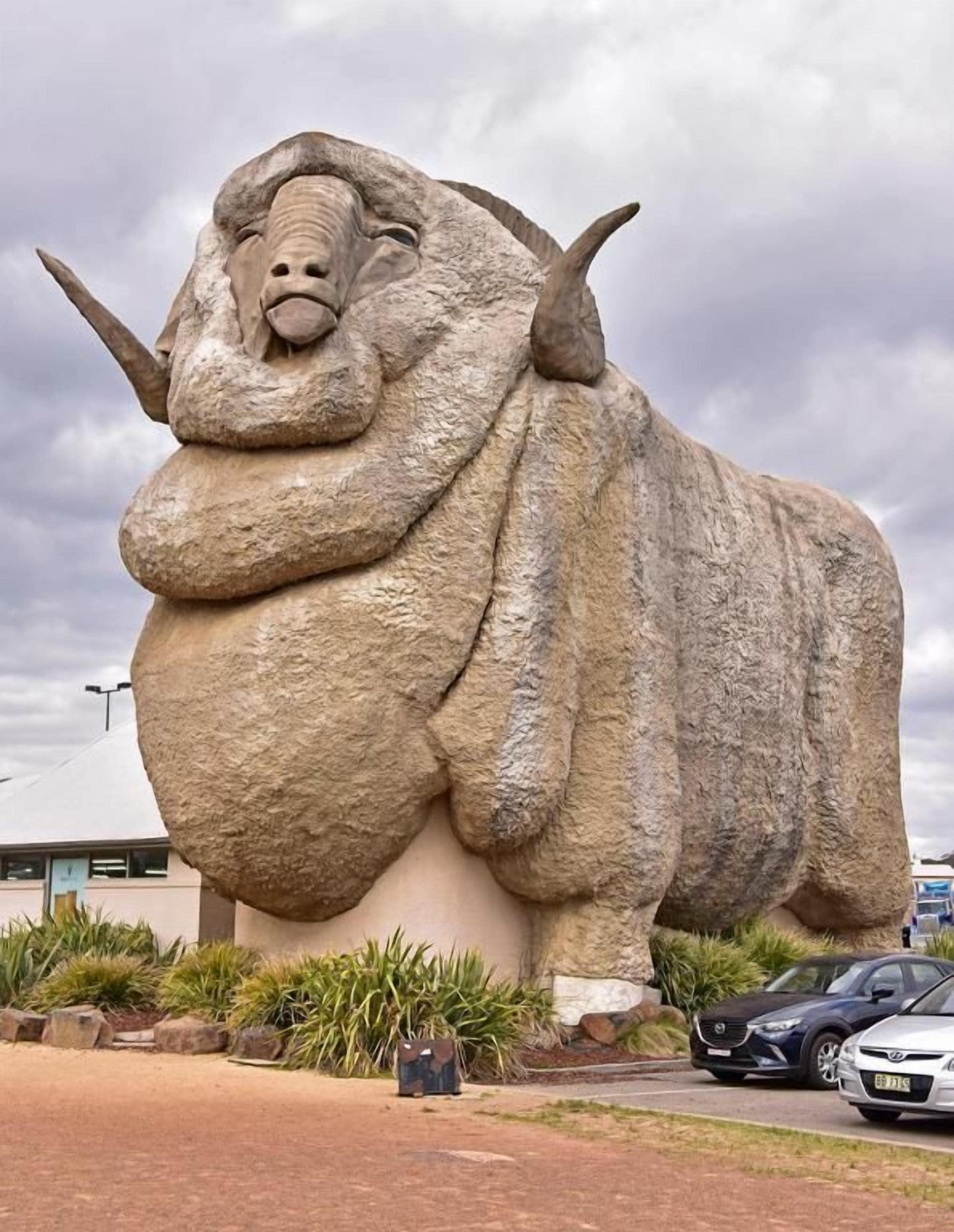 A large stone sculpture of a bull with horns, located outdoors with a building and cars in the background.