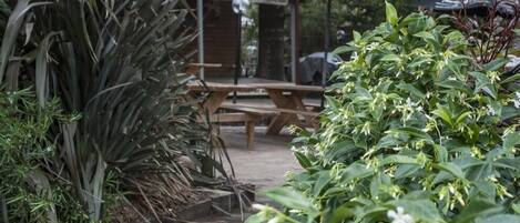 Un jardín con banco de madera y mesas de picnic bajo sombrillas, rodeado de plantas y follaje.