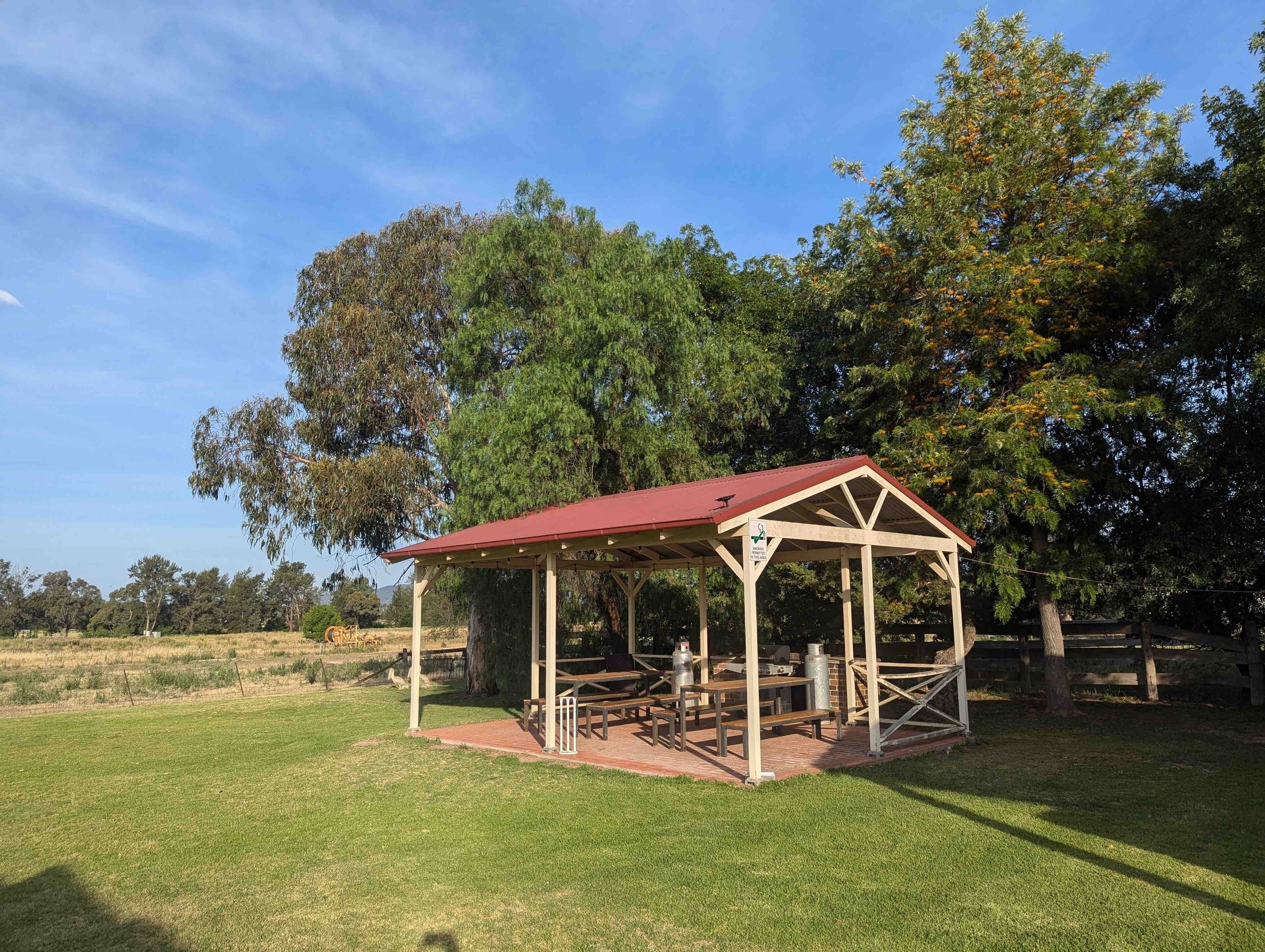 Een pergola met een rood dak en houten constructie, omgeven door gras en bomen.
