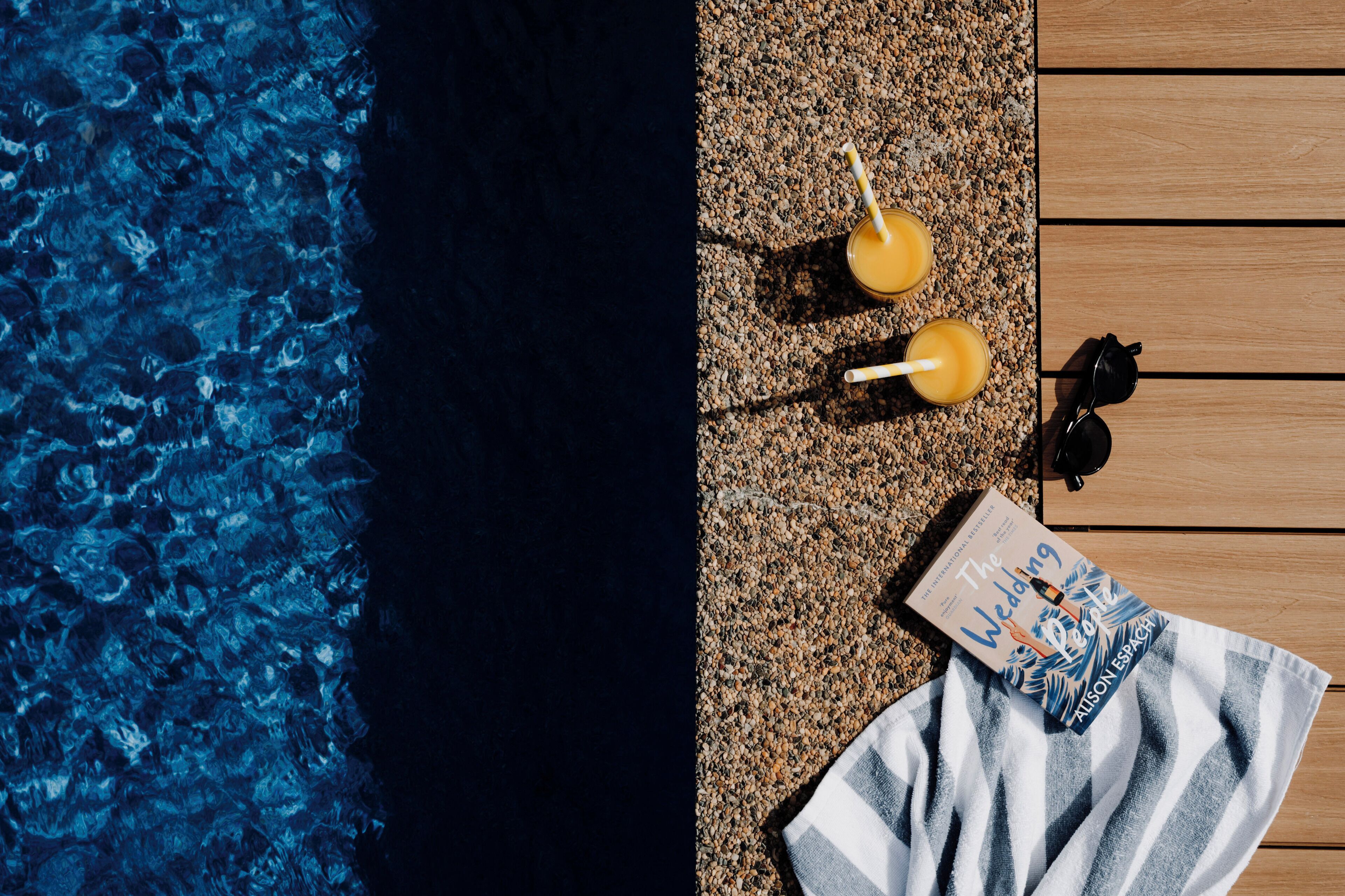 Un área junto a la piscina con un deck de madera, dos bebidas con pajitas, un par de gafas de sol y un libro titulado 