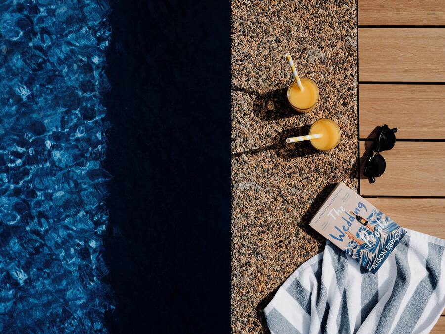 Un área junto a la piscina con un deck de madera, dos bebidas con pajitas, un par de gafas de sol y un libro titulado