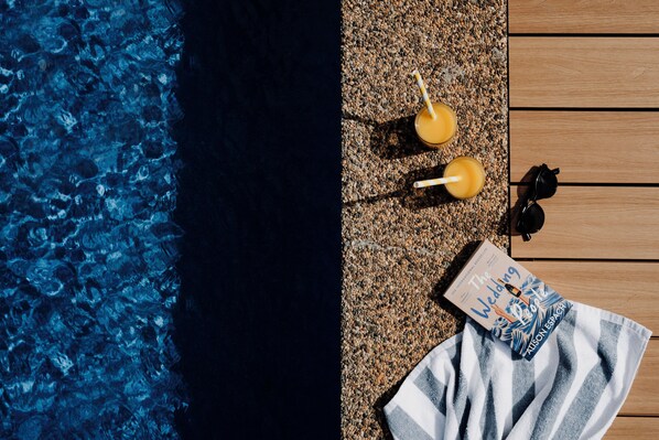 Un área junto a la piscina con un deck de madera, dos bebidas con pajitas, un par de gafas de sol y un libro titulado