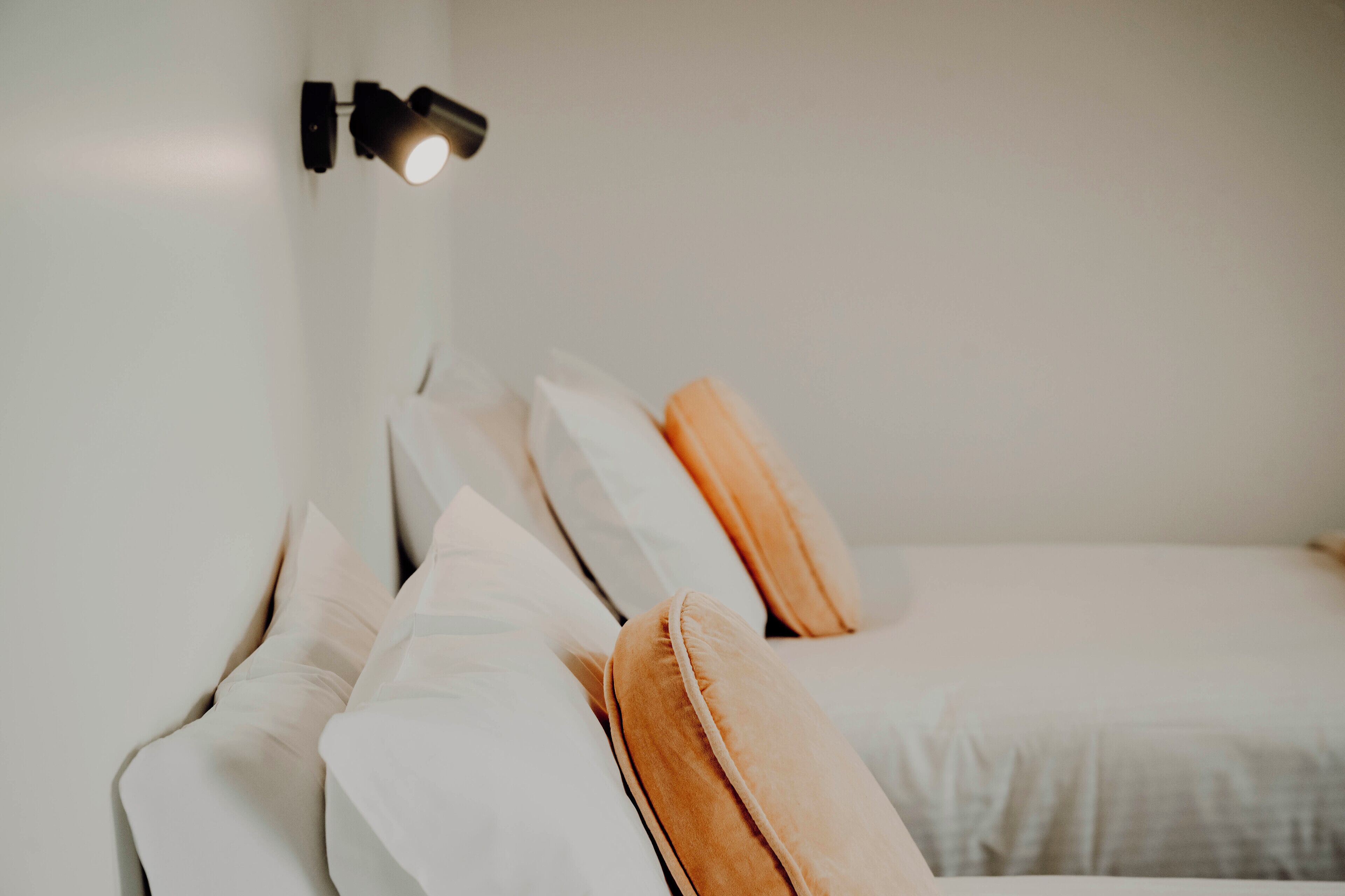 A neatly made bed with white and orange pillows, a black wall-mounted lamp, and a plain white wall.