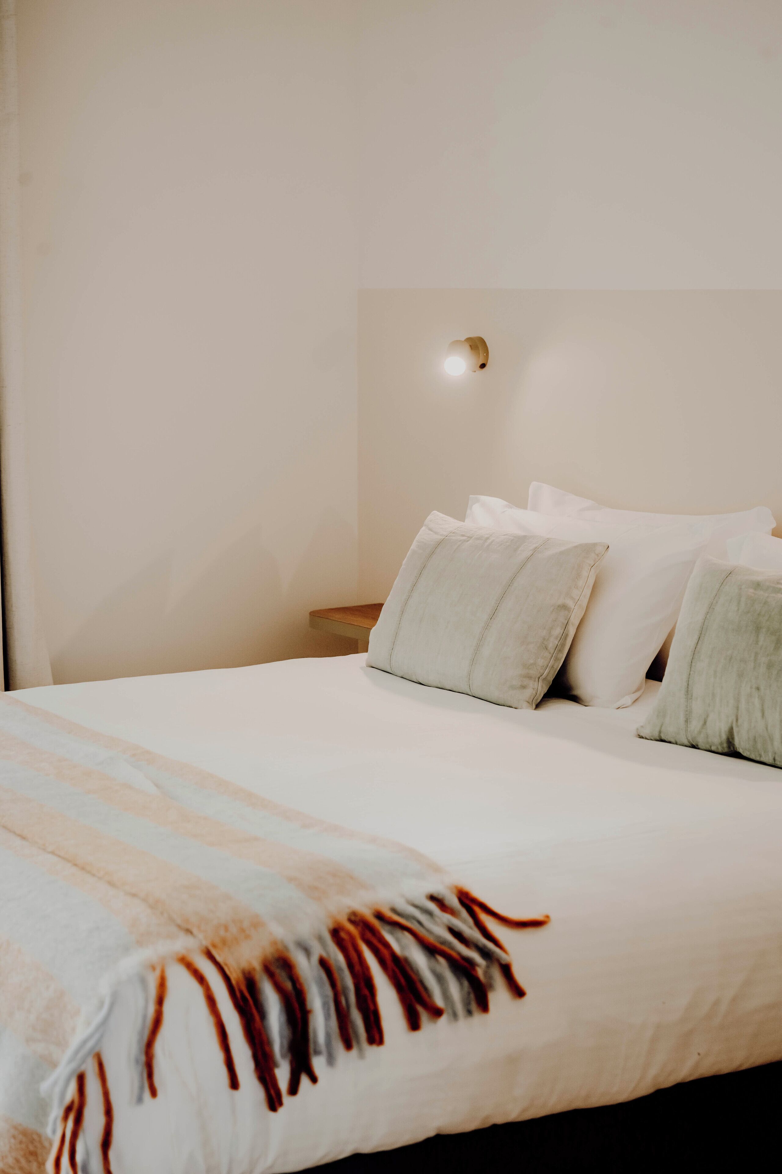 A neatly made bed with a striped blanket and pillows, a bedside table with a lamp, and a plain wall.