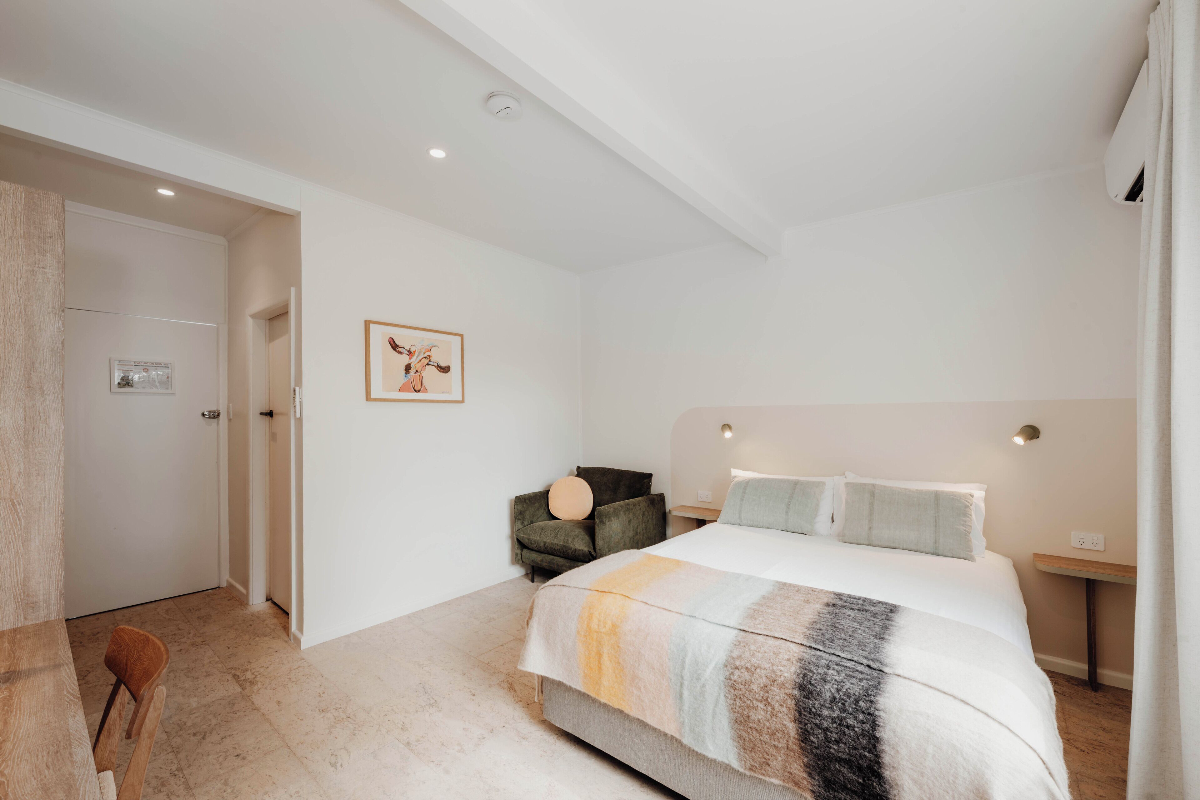 A bedroom with a bed, a chair, a bedside table, and a wall-mounted light fixture.