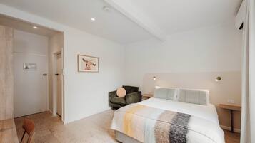 A bedroom with a bed, a chair, a bedside table, and a wall-mounted light fixture.
